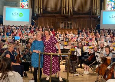 musicians sitting down on stage, with Angela Kay and Libby Croad standing up and posing for photo at the front